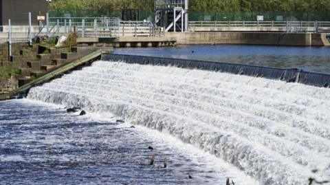 A river flows down a slope. A dam can be seen in the background.