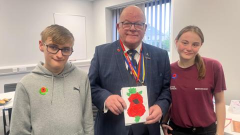 Mr Dean holds a hand made poppy card, and Matthew stands next to him on his left, and Rosie on the right.  All three smile for the camera.  The photo is taken in the classroom.