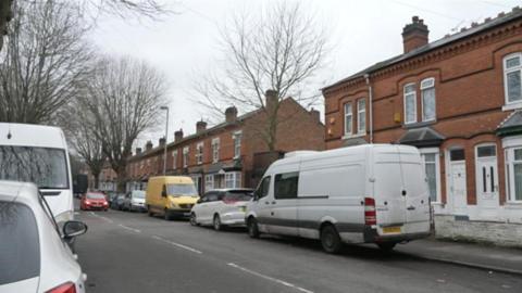 A street scene with vehicles parked either side of a road which is lined with terraced houses.