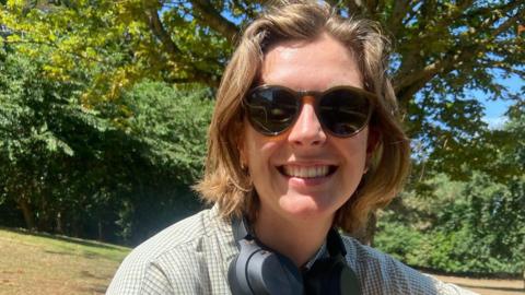Ms Faherty is smiling at the camera while outside in a field. Behind her is green grass and green trees. She has medium length light brown hair and is wearing sunglasses with headphones around her neck and a checkered white and green shirt.