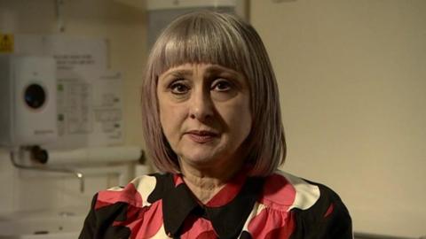 A woman with a short bob haircut, grey coloured with a slight purple tint, looks directly at the camera. She is not smiling. She wears a black, pink and white patterned blouse.