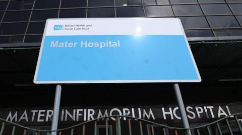 A blue hospital sign at the front of the Mater Hospital in Belfast.  The tall building has a dark glass facade.  The entrance also has a sign that says: "Mater Infirmorum Hospital" in large silver lettering.  The blue sign, erected by the Belfast Health and Social Care Trust, is behind a railing with chipped green paint. 