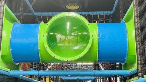 The image shows a section of a children's indoor playground. It shows a large cylindrical tunnel made of blue and green plastic, with a transparent green middle section. The tunnel is supported by blue padded bars and enclosed with safety netting. 