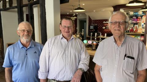 Three men, all wearing glasses and in summer shirts, stand in front of a pub bar with draught taps, bottles of alcohol and packets of crisps seen behind them.
