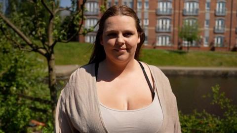 Louise stands in front a lake, she is wearing a beige vest top with beige cardigan. Her hair is dark and set back off her face.