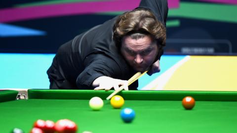 A snooker player with a black outfit and black headband cueing up the white ball from the baulk end of the table