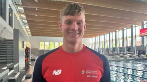 Ollie Morgan has light brown hair and is smiling at the camera. He is standing in front of an indoor swimming pool. He is wearing a red and black t-shirt with the University of Birmingham logo on it.