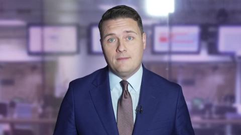 Wes Streeting, he has short dark hair, is clean shaven and is wearing a blue suit with white shirt and a metallic purple-grey colour tie