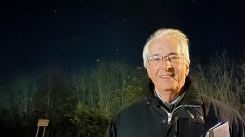 Stars in the night sky twinkle behind astronomer Nick Hewitt, who is wearing a black winter jacket and has grey hair and glasses. There is a notebook under one of his arms.