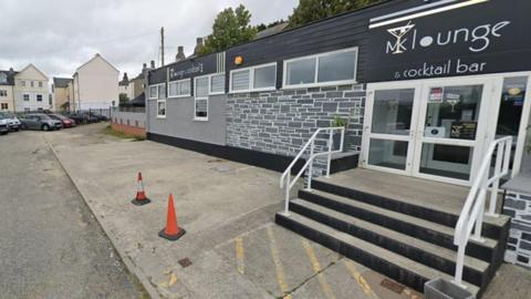 A street view of the Mk Lounge in Bodmin, Cornwall. There are four steps leading to the entrance which has two double white doors. Above the doors is a black sign which has the words, in white, 'MK lounge & cocktail bar'. The K is merged with a martini glass. To the left is the pavement with two orange traffic cones near the steps. There are houses in the background.