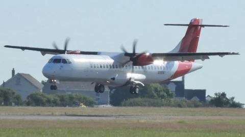 The ATR aircraft, G-PEMB, landing at Guernsey Airport. The white aircraft has red wings and tail and is about to touch down on the runway