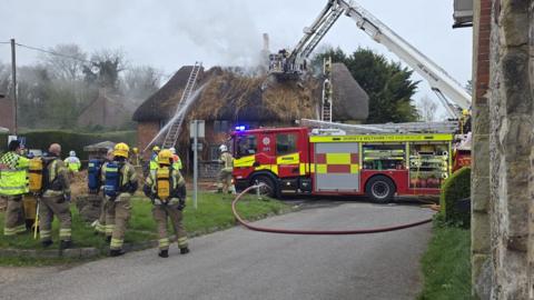 A fire engine with red and yellow hi-vis decals parked in the middle of the road. There are around nine firefighters standing nearby - one spraying water from a hose onto the thatched roof cottage, which is visible in the distance. Smoke can be seen coming from the destroyed roof, which appears burnt and pulled apart.