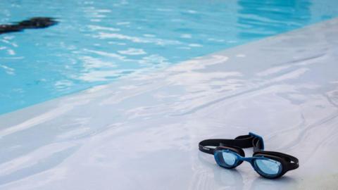 A stock image of a pool with some black and blue goggles lying on the side. 