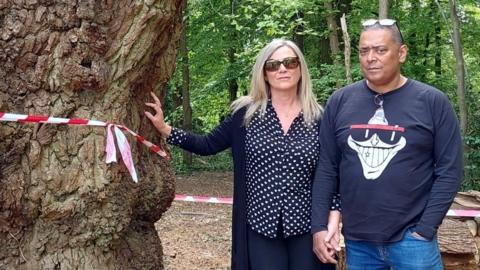 A man and a woman stand hand in hand next to a felled tree, the woman with her other hand touching its remaining stump.