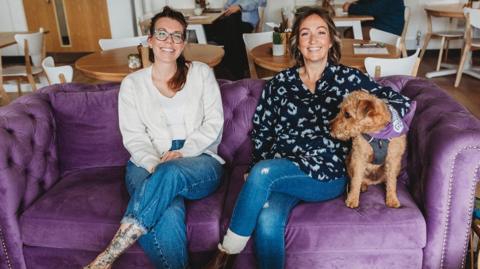 Kirsty, a trustee, and Sophie are sitting on a plush velvet couch in the cafe. Sophie (on the right) has a small, brown curly-haired dog sitting under her arm. Both are smiling broadly and have their legs crossed. Two people can be seen sitting at tables behind them. 