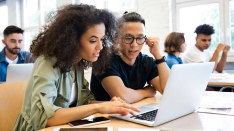 Stock photo of students conversing while using a laptop in a classroom with other students in the background