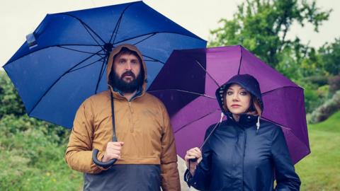 A man and a woman stand under umbrellas. They are both wearing rain jackets with the hoods up