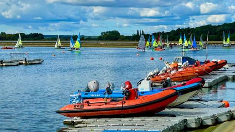 Colourful boats on Farmoor Reservoir near Cumnor. There are several orange RIBS on a floating  pontoon in the foreground. Behind there are sailing dinghys with colourful sails. The water is blue and the sky overhead is full of white clouds. On the horizon you can see a row of green trees.