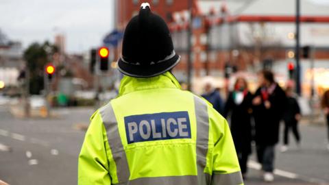 A British policeman, with his back to the camera and wearing a hi-vis police coat, watches people walking by across a road, which is blurred with him in focus.