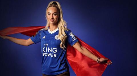 Alisha Lehmann in a Leicester shirt in front of a blue background and holding a red flag