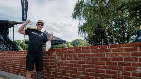 A man in a black tshirt and cap is standing next to a brick wall holding a trowl.