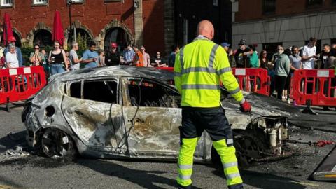 A crowd gather as a burned out car is towed away.