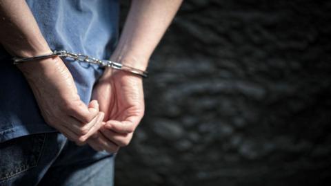 A close-up photo showing a pair of hands in handcuffs. The person's back can also be seen. They are wearing jeans and a blue T-shirt.