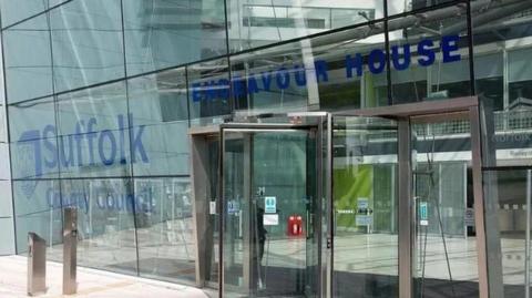 The glass fronted doors of Endeavour House of Suffolk County Council's main building in Ipswich. It has blue writing saying Suffolk County Council on it. 