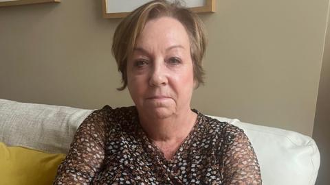 Deborah Crisp, a woman with short light brown hair wearing a brown patterned top. She sits on a white sofa next to a yellow cushion, in front of a beige wall.