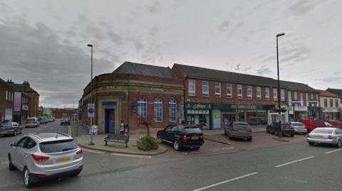 Northallerton High Street junction with Zetland Street, with pedestrians, shops and cars.