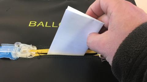 A hand holding a piece of paper and placing it into a box which says ballot box. 