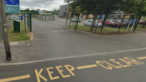 Part of a road with the words keep clear in yellow is at the bottom of the image. A sign that includes the words Elm Tree Primary Academy is on the left and buildings are visible in the background.