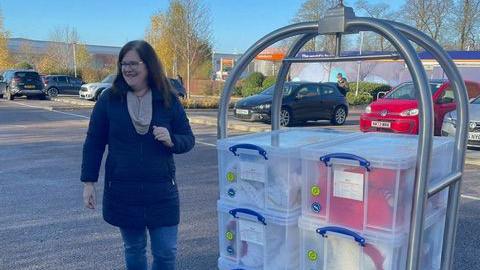 A woman is standing next to a trolley in a car park. There are plastic containers stacked on the trolley full of wool. 