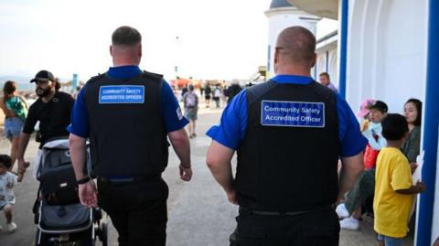 Two men walking wearing black uniform and a blue badge that reads community safety accredited officer. 