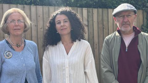 Left to right: Rosie Budd, Dr Stella Bolaki and Professor Stephen Banfield. Rosie is wearing a blue knit cardigan and has a short bob haircut. Dr Bolaki has long, dark curly hair and is wearing a white shirt, while Professor Banfield is wearing a maroon jumper, khaki jacket and flat cap. He is also wearing glasses.