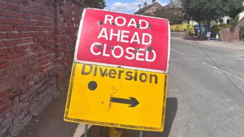 A bright red "road ahead closed" sign, on top of a bright yellow "diversion" sign on a road. There is a red brick wall on the left hand side of the signs, and there are a few roofs visible in the background.