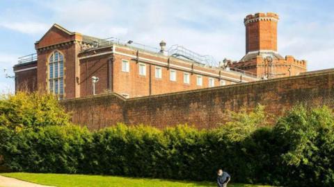 Red brick wall surrounds large building with long glass window, with multiple windows at height and a turret.