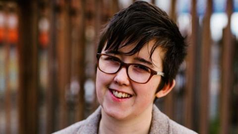 A smiling Lyra McKee standing in front of iron railings. She is wearing a brown jacket and eye glasses, and has short dark hair.