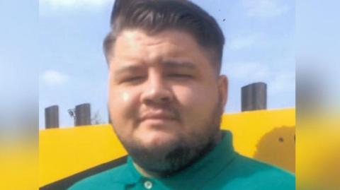 Euan Johnston, a young man with slicked back dark hair and a turquoise green polo shirt, looks into the camera. His eyes are squinting as the sun shines brightly and has a very slight smile. He is standing against a bright yellow wall.