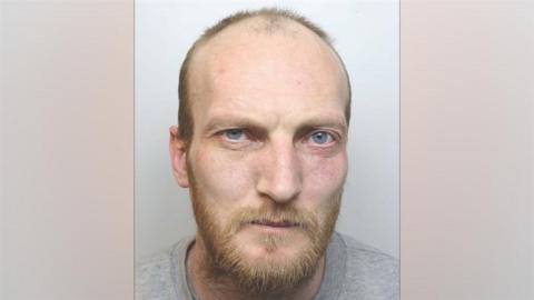 A police mugshot of John Whitfield, who has blue eyes, receding hair and a ginger beard