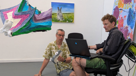 Dr Ben Kyneswood is on the right sat down on a chair staring at a computer screen while Derek Lawrence is on the left sat on the floor holding a grey and white projector. 