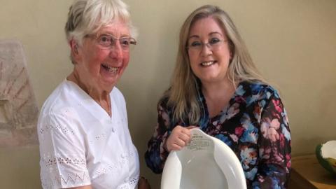 Mary Jacobs smiles while standing next to Julie Stevenson. Mary is looking away from the camera and is wearing a white blouse. She has short grey hair and is wearing glasses. Julie is smiling as she looks at the camera while holding a bedpan. She is wearing a floral blouse and has long blonde hair and glasses.