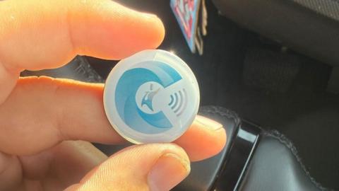 A close-up of a person's hand holding up a small, blue and white plastic disc about the size of a poker chip. They appear to be in a car with black interior. 
