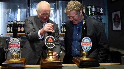 King Charles, wearing a grey suit, stands behind a bar and pulls a pint while a man wearing a black jacket and blue shirt stands next to him and watches.