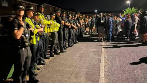 People are gathered in the street outside a property. A row of police officers are visible outside the house. Some of the people are wearing hoodies and holding mobile phones.