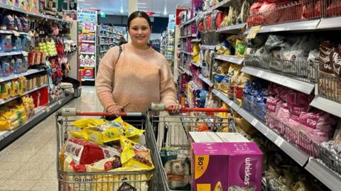 Tyesha Muncaster stands in the aisle of a supermarket. In front of her are two trolleys which are filled with pet food.