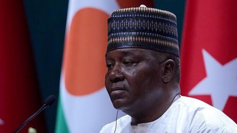 A head and shoulders image of Abdourahamane Tchiani with him looking to the left of the picture. He is wearing a traditional cap and, out of focus, the flags of Niger and Turkey can be seen behind him
