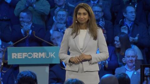 Suella Braverman in a grey and white suit beside a speaking lectern with Reform UK to her left. Audience is visible behind her lit by blue-purple lighting.