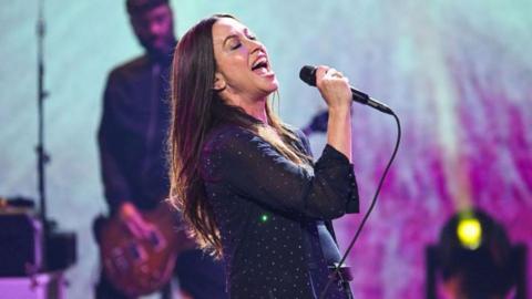 Alanis Morissette, wearing a sparkly black blazer and jeans with a black belt, singing into a microphone with her long dark hair behind her as she leans back. She is on a stage with lights and a guitarist behind her.