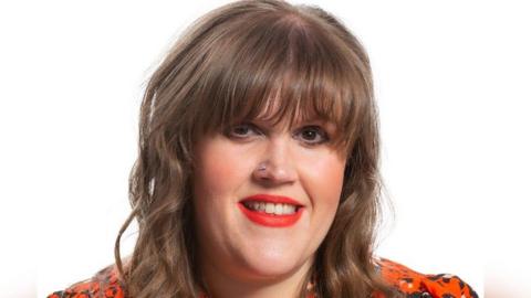 Eleanor Wills, who has brown hair, smiles wearing an orange and black blouse for an official council photograph. 